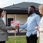 A couple shaking hands with a real estate agent in front of a "Sold" sign.