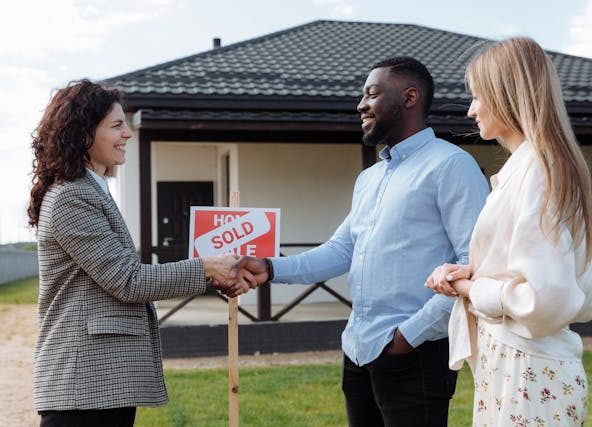 A couple shaking hands with a real estate agent in front of a "Sold" sign.